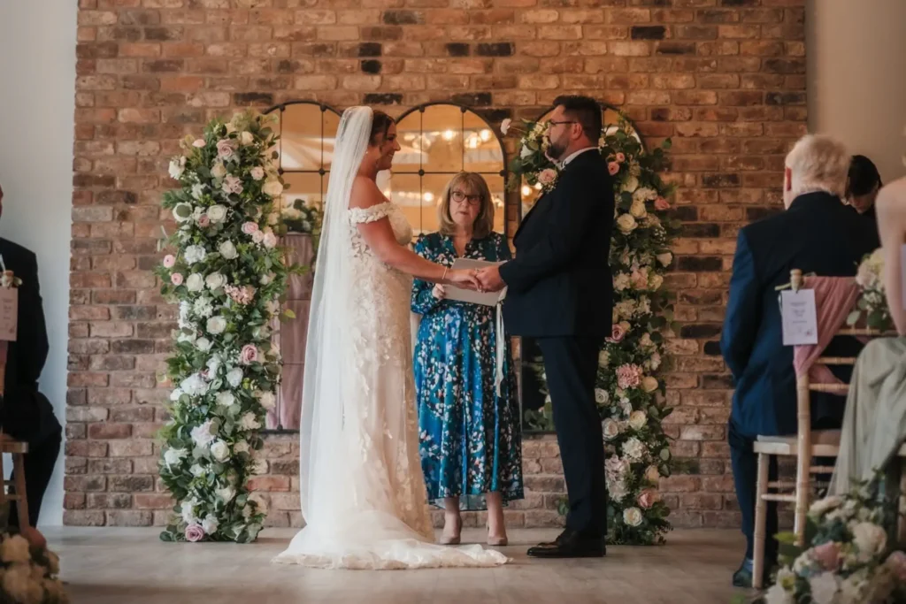 Bride and groom holding hands as celebrant Julie Goodall leads their indoor wedding ceremony beneath a floral arch